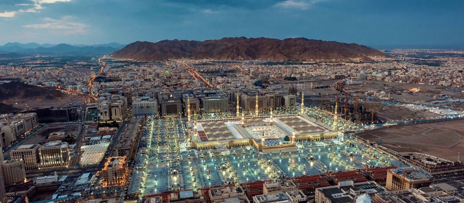 Madinah from the air at dusk: the Prophet's Mosque and its lit courtyards at the centre of the city, the mountains beyond.