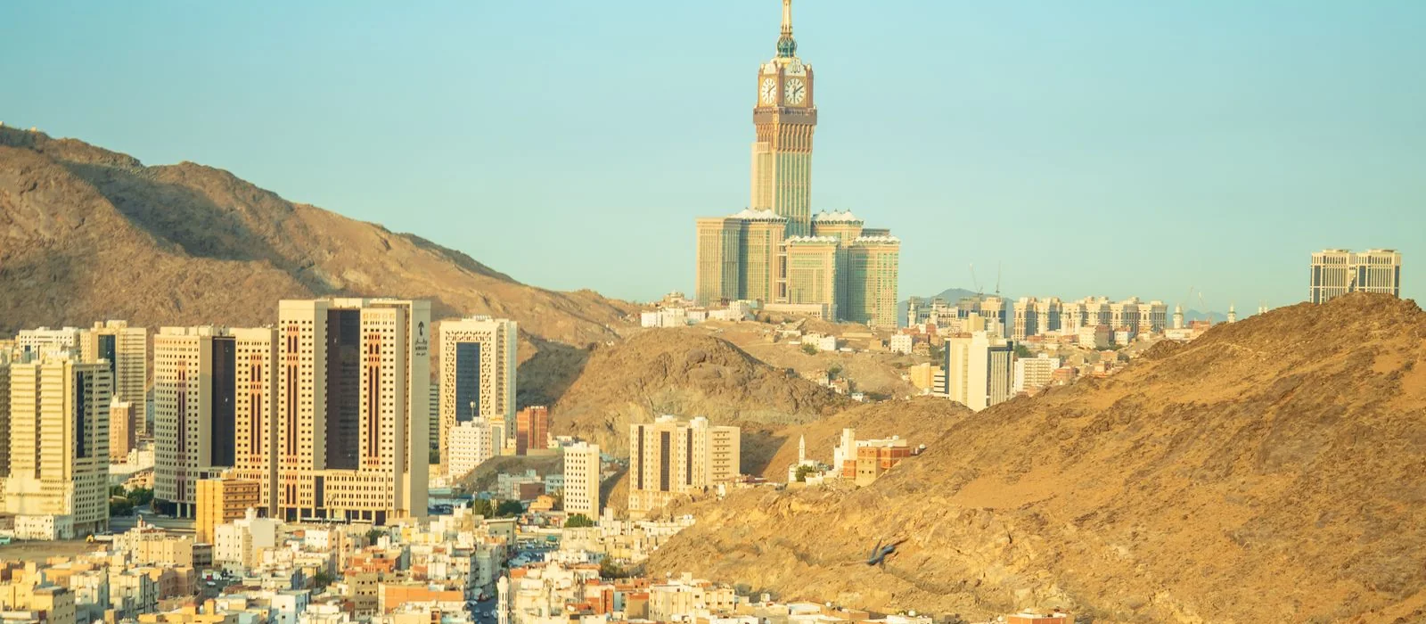 Makkah in the late afternoon: modern residential towers and dense housing filling the valley, the Abraj Al Bait clock tower rising beyond the golden hills.